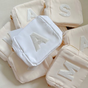 May include: A collection of small, cream-colored cosmetic bags with gold zippers. Each bag features a large, embellished letter in silver beads. One bag is white. The letters include A, S, and M.