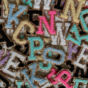May include: A collection of colorful, embroidered fabric letters with gold glitter trim. The letters are in various colors, including pink, blue, white, black, and yellow. The letters are in a variety of fonts and sizes.