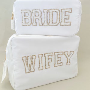 May include: Two white zippered cosmetic bags with gold glitter lettering. The top bag says "BRIDE" and the bottom bag says "WIFEY".
