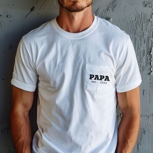 May include: White t-shirt with a pocket on the left side. The pocket has black text that reads "PAPA est. 2024".
