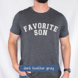May include: A dark heather gray t-shirt with the text "FAVORITE SON" in white.