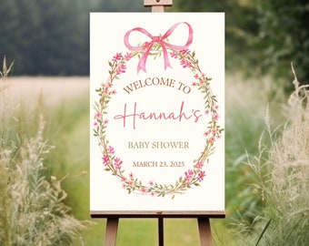 May include: A white welcome sign with a pink floral wreath and a pink bow. The sign says "Welcome to Hannah's Baby Shower March 23, 2025".
