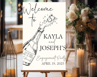 May include: Black and white welcome sign with a champagne bottle popping open. The sign reads "Welcome to Kayla and Joseph's Engagement Party April 19, 2025".