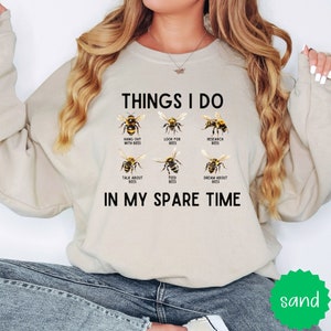 May include: A beige sweatshirt with the text "THINGS I DO" and a list of bee-related activities, including "hang out with bees", "look for bees", "research bees", "talk about bees", "feed bees", and "dream about bees". The text "IN MY SPARE TIME" is below the list.