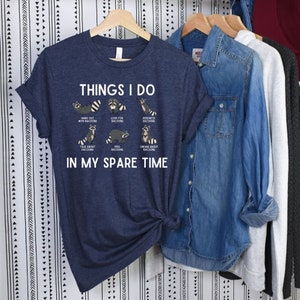 May include: A navy blue t-shirt with a white graphic of raccoons and the text "Things I do in my spare time". The graphic shows raccoons hanging out, looking for raccoons, researching raccoons, talking about raccoons, feeding raccoons, and dreaming about raccoons.