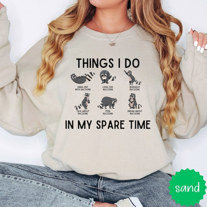 May include: A beige sweatshirt with a graphic print of raccoons doing various activities, including hanging out, looking for, researching, talking about, feeding, and dreaming about raccoons. The text reads "Things I do in my spare time".