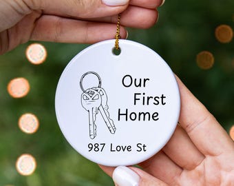 May include: A white, round ceramic ornament with a gold string for hanging. The ornament features a black line drawing of keys and the text "Our First Home" and "987 Love St."