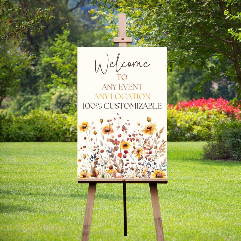 May include: A wooden easel displays a welcome sign with floral artwork. The sign reads "Welcome to Any Event Any Location 100% Customizable." The artwork features yellow sunflowers and other wildflowers in shades of brown, red, and orange.