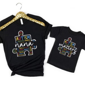 May include: Two black t-shirts with a puzzle piece design. The larger shirt says "Autism Awareness Aware nana" and the smaller shirt says "Autism Matters". Both shirts have colorful text around the puzzle piece.