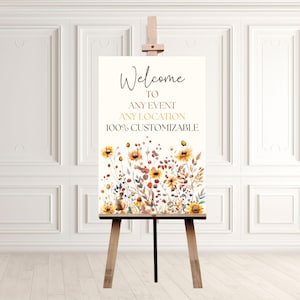 May include: A wooden easel displays a welcome sign with floral artwork. The sign reads "Welcome to Any Event Any Location 100% Customizable." The floral design features yellow, orange, and red flowers with green and brown foliage.