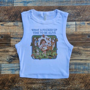 Op de afbeelding: Een wit mouwloos crop-top shirt met een grafische print van een meisje in een jurk in een bos met een konijn, een hert, vogels en vlinders. De tekst boven de illustratie luidt: "WHAT A FUCKED UP TIME TO BE ALIVE" in blauwe letters.