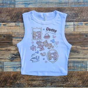 May include: A light blue sleeveless crop top with a graphic print of Duffy and Friends. The print features the characters Duffy and ShellieMay in various poses, along with hearts, balloons, and other cute details.