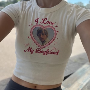 May include: Cream crop top with the text "I Love My Boyfriend" in pink script. The shirt features a heart-shaped graphic with a portrait of a man. The top is paired with black leggings.