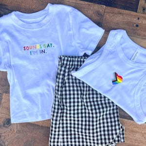 Puede incluir: Una camiseta blanca de manga corta con el texto bordado SOUNDS GAY. I'M IN. en colores arcoíris, una camiseta de tirantes blanca con un pequeño corazón arcoíris bordado y unos pantalones cortos con estampado de cuadros vichy.