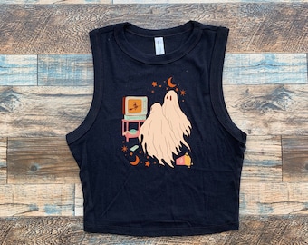 May include: A black sleeveless crop top featuring a graphic print of two pale ghosts watching a vintage television set. The illustration includes a small witch on the screen, orange stars, a crescent moon, and a container of popcorn on the floor.