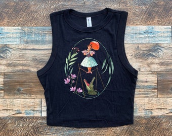 May include: Black sleeveless crop top with a graphic print of a woman standing on a scale with an alligator below. The woman is holding an umbrella and is surrounded by flowers and leaves.