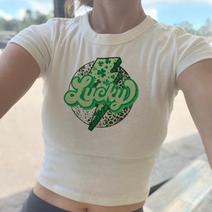 May include: White crop top with a green and black graphic of a lightning bolt with the word "Lucky" in a retro font. The graphic is surrounded by shamrocks.