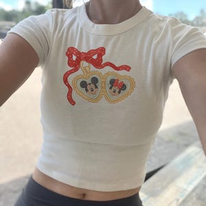 Op de afbeelding: Crèmekleurige crop top met een Mickey en Minnie Mouse afbeelding in hartvormige kaders. Een rode strik met witte stippen siert de top. Het shirt heeft korte mouwen en een ronde halslijn.