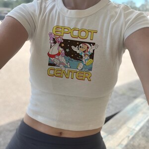 Puede incluir: Top corto color crema con las palabras "EPCOT CENTER" en letras amarillas y rojas. El gráfico presenta personajes de dibujos animados con trajes espaciales. La parte superior es de manga corta y tiene cuello redondo.