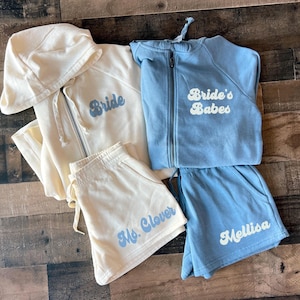 Puede incluir: Dos conjuntos a juego de sudaderas con capucha y pantalones cortos de color crema y azul claro. El conjunto crema tiene la palabra "Bride" en la sudadera y "Ms. Clover" en los pantalones cortos. El conjunto azul claro tiene las palabras "Bride's Babes" en la sudadera y "Melissa" en los pantalones cortos.