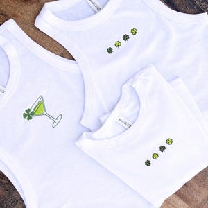 May include: White tank tops and t-shirts with St. Patrick's Day designs. One tank top features a green cocktail with a shamrock, while others have embroidered shamrocks. The shirts are displayed on a wooden surface.