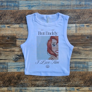 May include: A light blue sleeveless crop top with a graphic print of a cartoon mermaid with red hair. The text "EST 1989 But Daddy I Love Him" is printed on the shirt.