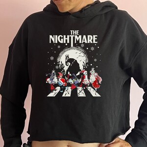 Puede incluir: Sudadera corta negra con un gr&aacute;fico de "Pesadilla antes de Navidad". El dise&ntilde;o incluye el texto "THE NIGHTMARE" sobre una escena de personajes cruzando un paso de cebra frente a una luna llena.