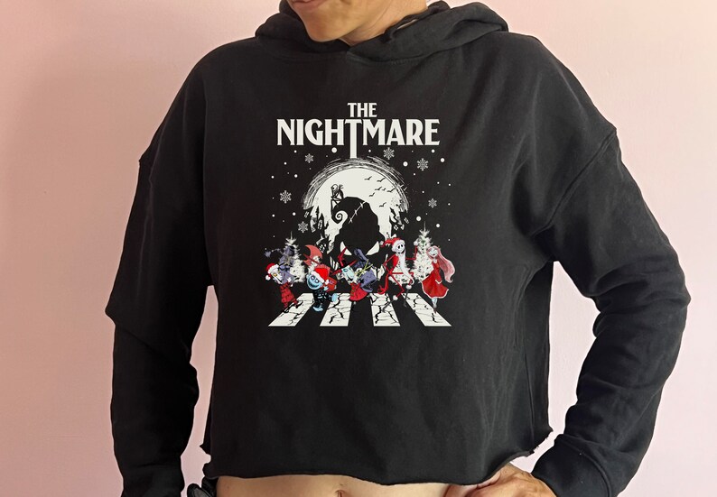 Puede incluir: Sudadera corta negra con un gr&aacute;fico de "Pesadilla antes de Navidad". El dise&ntilde;o incluye el texto "THE NIGHTMARE" sobre una escena de personajes cruzando un paso de cebra frente a una luna llena.