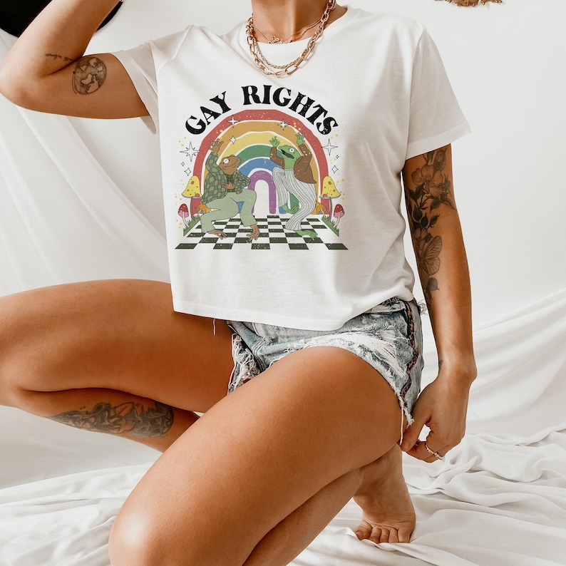 May include: White t-shirt with the words "GAY RIGHTS" above a rainbow and two cartoon frogs dancing on a checkered floor. The shirt has a retro, whimsical design with mushrooms and stars.