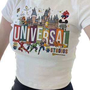 May include: White t-shirt with a colorful graphic print of the Universal Studios logo, featuring characters like Spiderman, the Simpsons, and Minions.