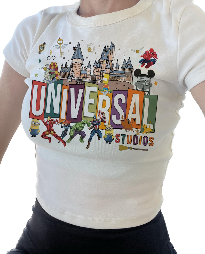 May include: White t-shirt with a colorful graphic print of the Universal Studios logo, featuring characters like Spiderman, the Simpsons, and Minions.