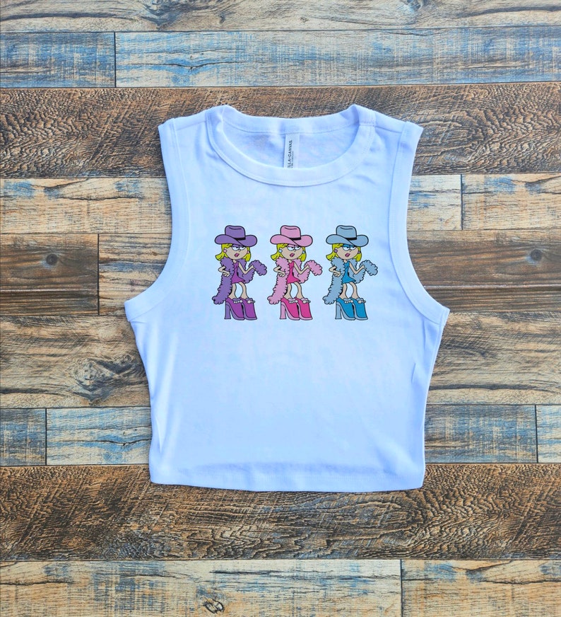Op de afbeelding: Een witte mouwloze crop top met een herhalende grafische print van een stripfiguur met een cowboyhoed, een veren boa en plateauschoenen. Het personage is afgebeeld in drie verschillende kleurvariaties: paars, roze en blauw.