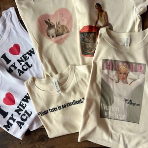 May include: Five different t-shirts with various designs. One white tank top with a red heart and the text "I love my new ACL". Another white tank top with a red heart and the text "I love my new ACL". A cream colored t-shirt with a pink heart and two brown rabbits. A cream colored t-shirt with the text "Your taste is so excellent." A cream colored t-shirt with a magazine cover featuring a woman and the text "Glamour Women of the Year Hannah Waddingham".