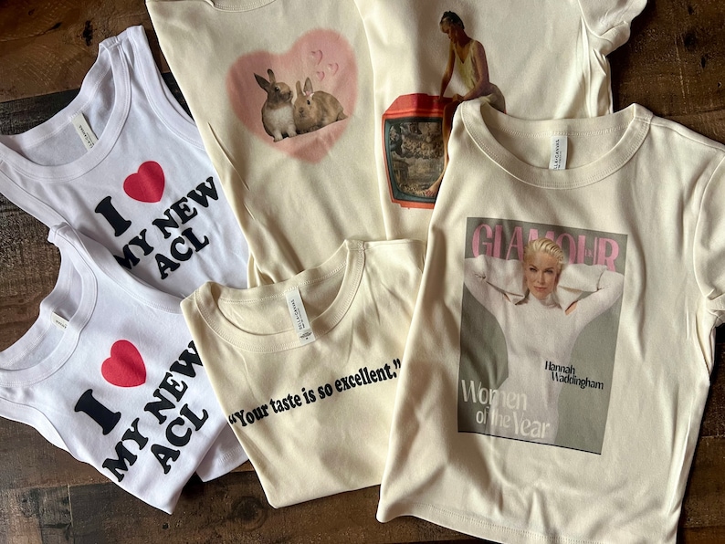 May include: Five different t-shirts with various designs. One white tank top with a red heart and the text "I love my new ACL". Another white tank top with a red heart and the text "I love my new ACL". A cream colored t-shirt with a pink heart and two brown rabbits. A cream colored t-shirt with the text "Your taste is so excellent." A cream colored t-shirt with a magazine cover featuring a woman and the text "Glamour Women of the Year Hannah Waddingham".