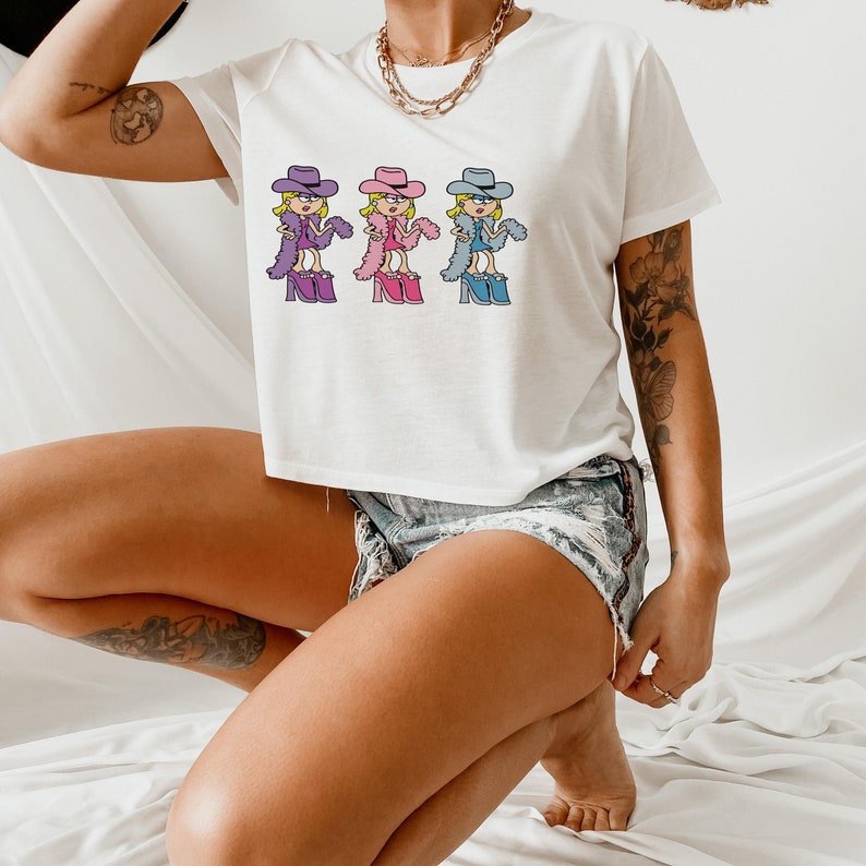 Op de afbeelding: Een wit T-shirt met korte mouwen en een grafische print van drie stripfiguren met cowboyhoeden, jurken en plateauschoenen. De figuren zijn gekleurd in paars, roze en blauw en dragen elk een bijpassende veren boa.