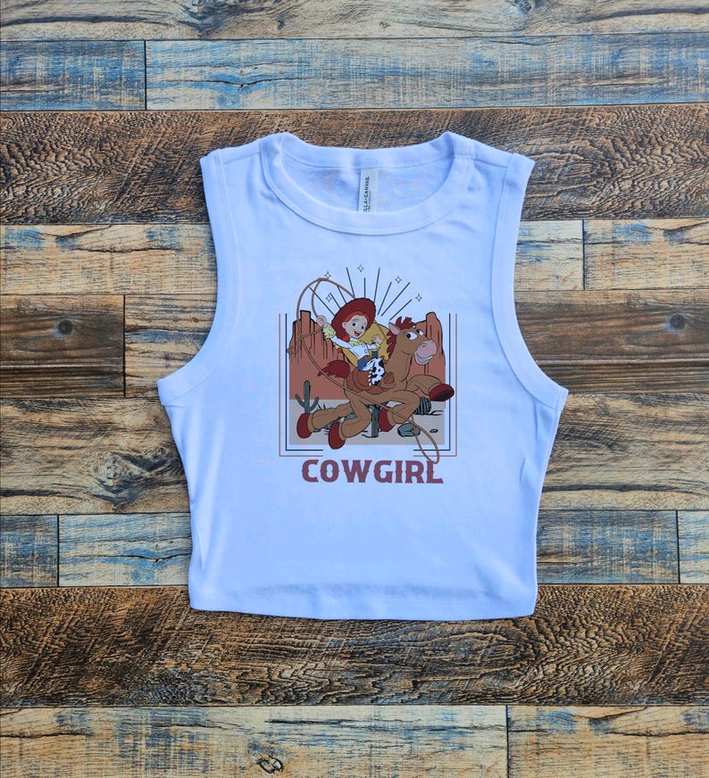 May include: White sleeveless crop top with a graphic print of a cartoon cowgirl riding a horse. The graphic is in a retro style with a red, orange, and brown color scheme. The text "COWGIRL" is printed below the graphic.
