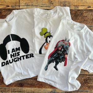 May include: Three white t-shirts with different designs. One shirt has a black and white graphic of a Star Wars character with the text "I am his daughter". Another shirt has a cartoon image of a character from Disney's Goofy movie. The third shirt has a graphic of Thor from Marvel Comics.