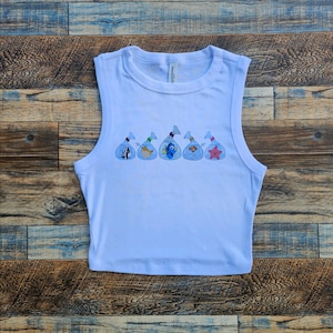 Pode incluir: Um top curto sem mangas azul claro com imagens bordadas de peixes em sacos plásticos. Os peixes incluem um peixe preto e branco, um peixe amarelo, um peixe azul, um peixe laranja e uma estrela do mar rosa.