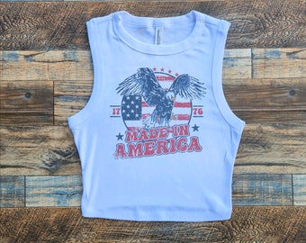 May include: White sleeveless crop top with a vintage-style graphic of an eagle flying over an American flag with the text "Made in America" and the years "1776" and "1776" on either side of the flag.