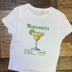 May include: White t-shirt with a green and white graphic of a margarita cocktail. The graphic includes the text "MARGARITA" and "Serve Me Ice Cold". The recipe for the cocktail is listed below the graphic: 50ml Tequila, 20ml Triple Sec, 25ml Lime Juice, Salt, Lime Wedge & Ice.