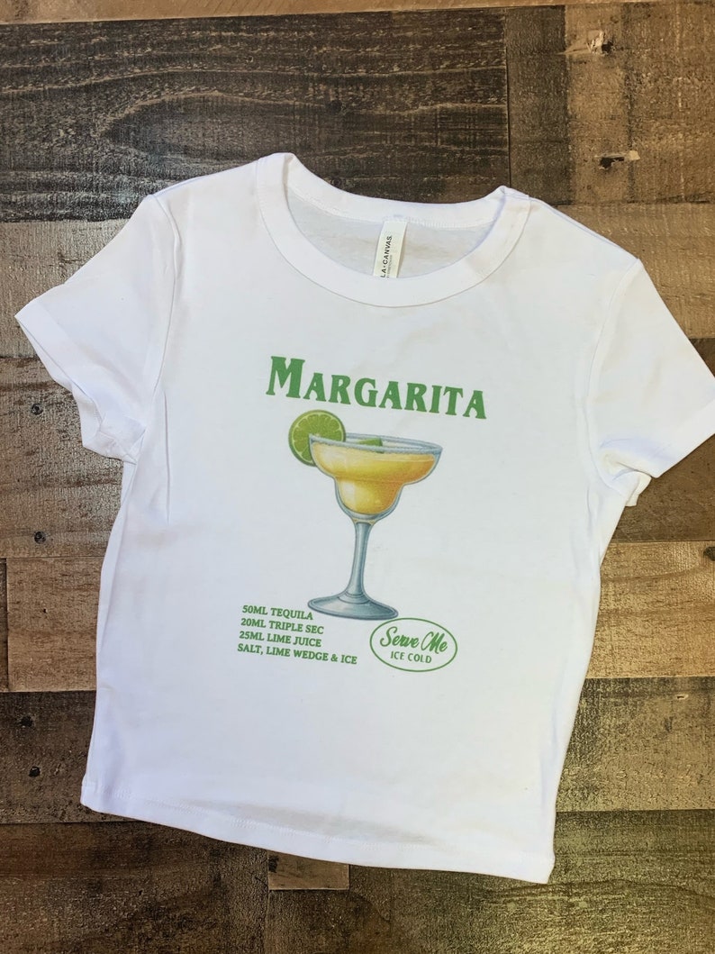 May include: White t-shirt with a green and white graphic of a margarita cocktail. The graphic includes the text "MARGARITA" and "Serve Me Ice Cold". The recipe for the cocktail is listed below the graphic: 50ml Tequila, 20ml Triple Sec, 25ml Lime Juice, Salt, Lime Wedge & Ice.