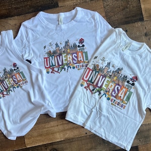May include: Three white t-shirts with a colorful graphic of Universal Studios landmarks and characters. The shirts are in different sizes and styles, including a tank top and two t-shirts.