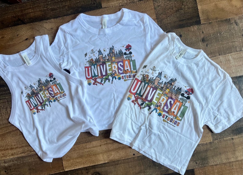 May include: Three white t-shirts with a colorful graphic of Universal Studios landmarks and characters. The shirts are in different sizes and styles, including a tank top and two t-shirts.