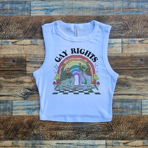 May include: White sleeveless crop top with a graphic print of two frogs dancing under a rainbow with the text "Gay Rights".