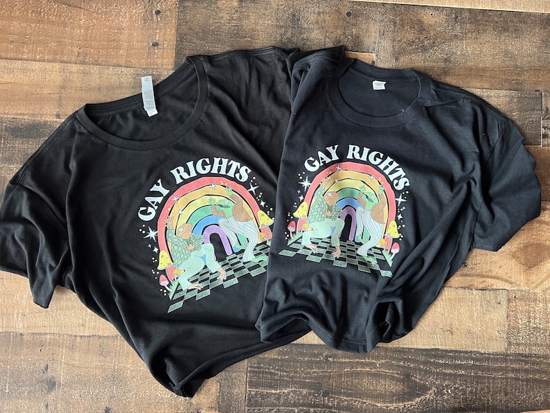 May include: Two black t-shirts with the words "GAY RIGHTS" in a rainbow design. The graphic features two figures, a rainbow, and a checkered floor. The shirts are casual and likely made of cotton.