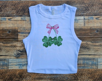 May include: A light blue sleeveless crop top with a pink bow and two green four-leaf clovers printed on the front.