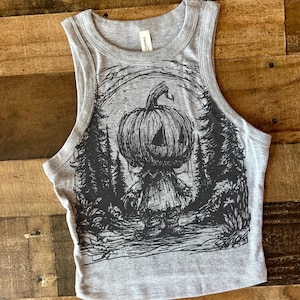 Op de afbeelding: Een grijze crop top met een zwarte afbeelding van een pompoenhoofdige figuur in een bos. De mouwloze top heeft een ronde hals en is gemaakt van een zacht materiaal. De afbeelding is een gedetailleerde illustratie met een Halloween-thema.