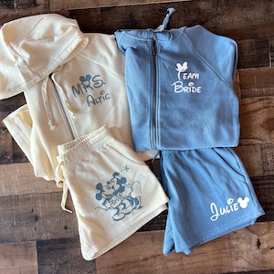 May include: Two matching sets of cream and blue hooded sweatshirts and shorts. The cream set has the text "MRS. Atric" and a Mickey Mouse couple graphic. The blue set has the text "Team Bride" and a Mickey Mouse head graphic. The blue shorts have the name "Julie" written on them.
