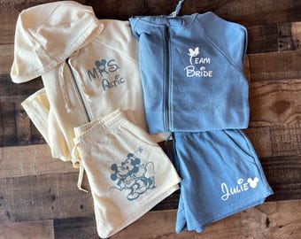 May include: Two matching sets of cream and blue hooded sweatshirts and shorts. The cream set has the text "MRS. Atric" and a Mickey Mouse couple graphic. The blue set has the text "Team Bride" and a Mickey Mouse head graphic. The blue shorts have the name "Julie" written on them.