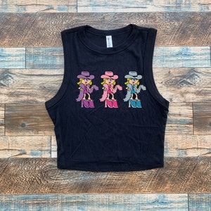 Op de afbeelding: Een zwarte mouwloze crop top met een grafische print van drie stripfiguren die cowboyhoeden, boa's en plateauschoenen dragen. De figuren zijn uitgevoerd in paarse, roze en blauwe kleurenschema's en horizontaal over de borst geplaatst.